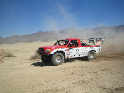 1990 Ford Bronco - red and white off-road race truck slides through a dirt corner, kicking up dust on a desert race course.