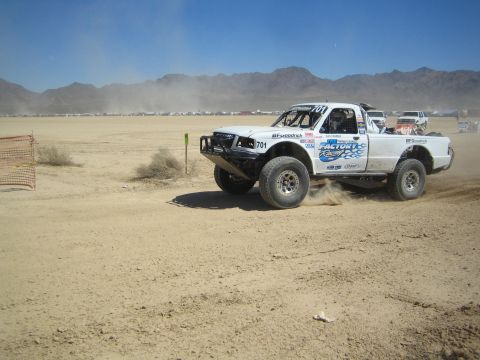 1990 Ford Bronco - white off-road race truck with Factory Racing graphics and number 701 races across a dusty desert flat.