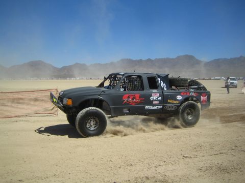 1990 Ford Bronco - black off-road race truck with GL Racing livery and number 702 slides through dirt on a desert race course.