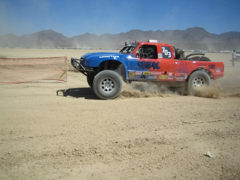 1990 Ford Bronco - red and blue off-road race truck marked number 703 slides through dirt, kicking up dust on a desert race course.