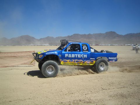 1990 Ford Bronco - blue off-road race truck with flame graphics and 'FABTECH' lettering, numbered 704, kicks up dust speeding across a desert course toward di…
