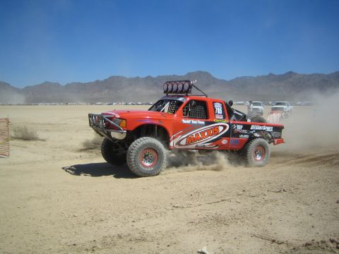 1990 Ford Bronco - red and orange off-road race truck with Maxxis sponsorship and roof light bar throws up dust racing across a dry desert flat under blue sky.