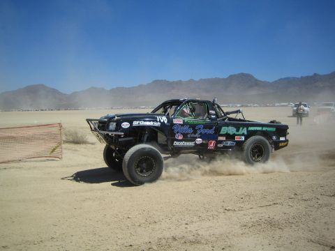 1990 Ford Bronco - black off-road race truck covered in sponsor decals including BFGoodrich kicks up a trail of dust racing across the desert toward mountains.