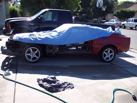 1971 Ford Mustang - A red car project is covered by a blue tarp, parked next to a black pickup truck.