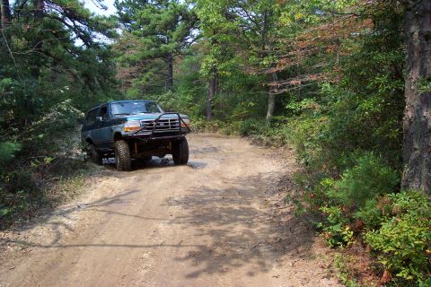 1987 Ford Bronco - A dark blue Ford Bronco navigates a dirt road through a wooded area.
