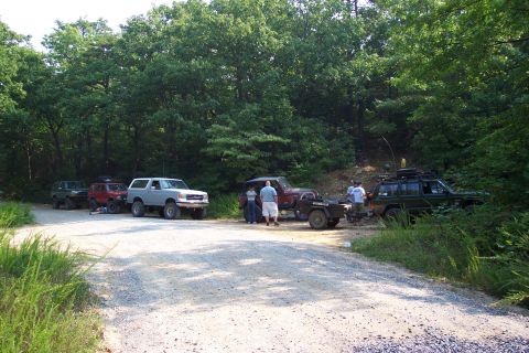 1987 Ford Bronco - Several off-road vehicles, including a 1987 Ford Bronco, are parked on a dirt road surrounded by trees.