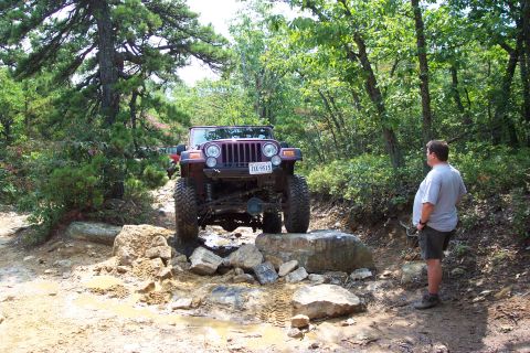 1987 Ford Bronco - A Jeep Wrangler TJ navigates a rocky obstacle on an off-road trail with a bystander observing.