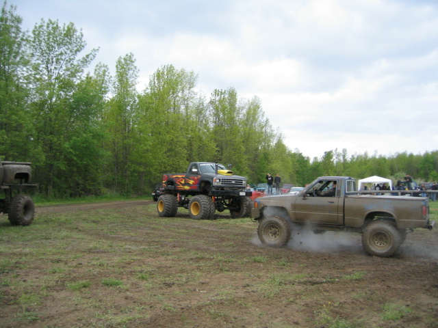 Muddy pickup truck kicking up dust, monster truck in background
