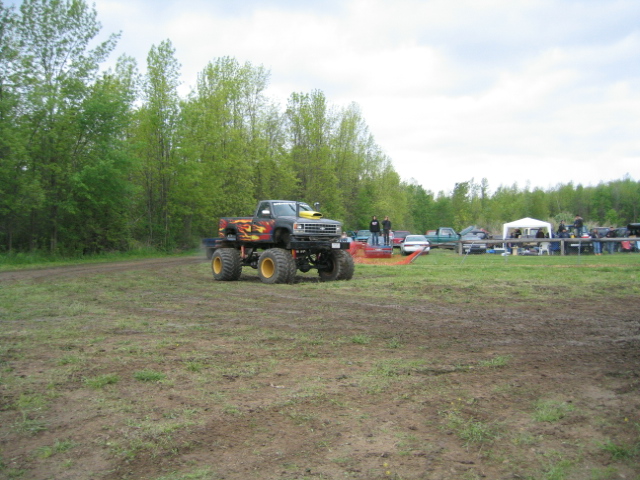 Monster truck with large tires and flame decals on a dirt field, with other vehicles and tents in the background