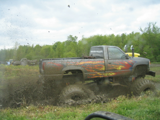 Lifted pickup truck with custom graphics driving through mud, kicking up spray. Another truck is visible in the background