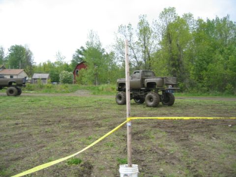1978 Ford Bronco - Two monster trucks are visible in a muddy field with trees and buildings in the background.