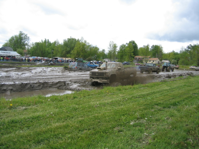Mud bogging event with several trucks in the mud, spectators in the background