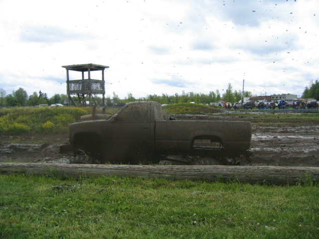 Mud-covered pickup truck stuck in mud pit with a wooden observation tower in background