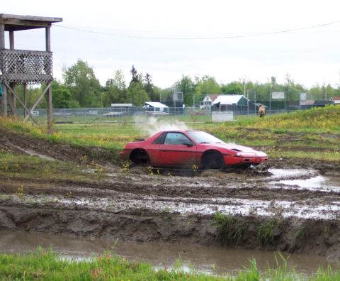 1978 Ford Bronco - A red Pontiac Fiero is stuck in a mud pit, kicking up dust and smoke.