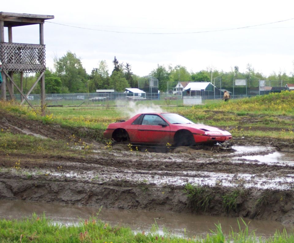 Red Pontiac Fiero stuck in mud pit with smoke coming from wheels