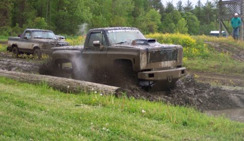1978 Ford Bronco - Two pickup trucks navigate a muddy obstacle course, kicking up dirt and water.