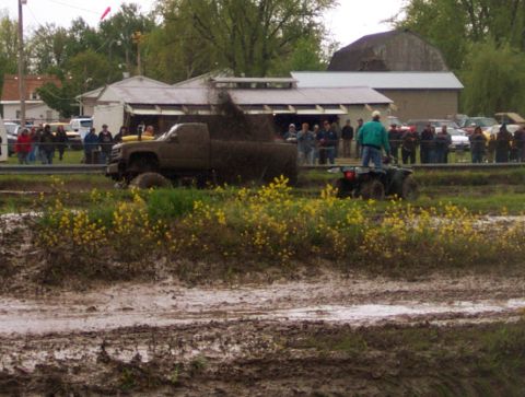 1978 Ford Bronco - A pickup truck churns up mud during a bogging event, with spectators and an ATV in the background.
