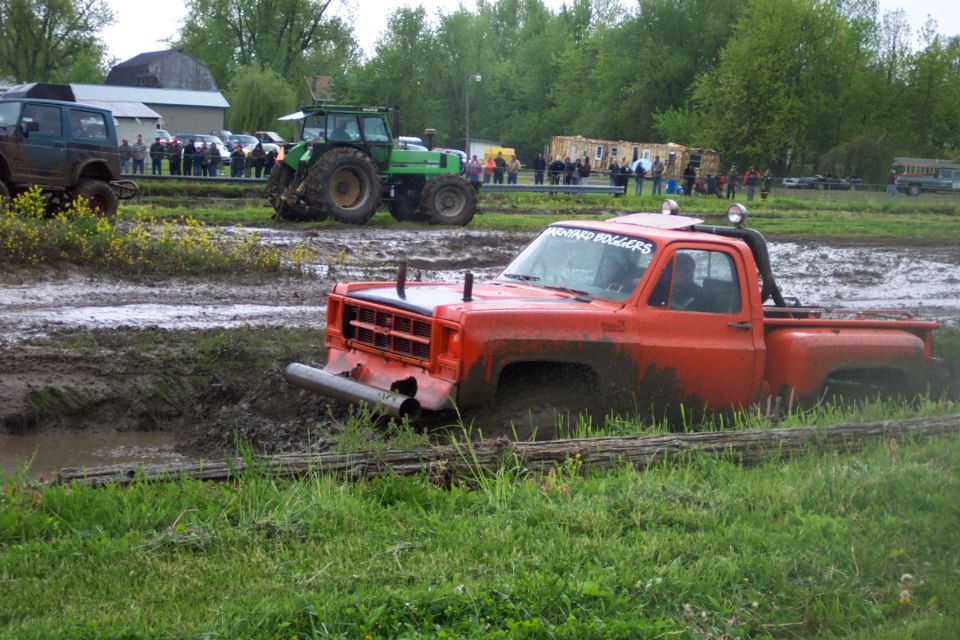 Orange pickup truck stuck in mud with a green tractor and other vehicles in the background