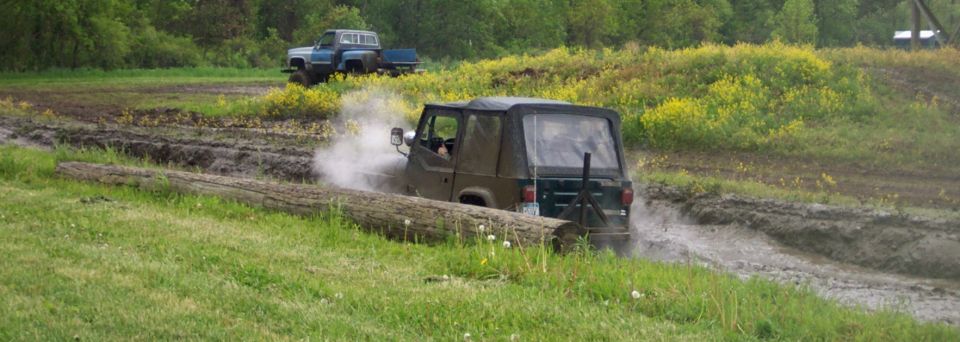 Dark off-road vehicle splashing through mud, with a blue pickup truck in the background