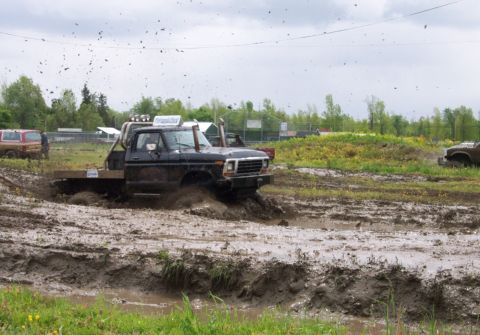 1978 Ford Bronco - Multiple trucks navigate a muddy track during an event, kicking up mud and debris.