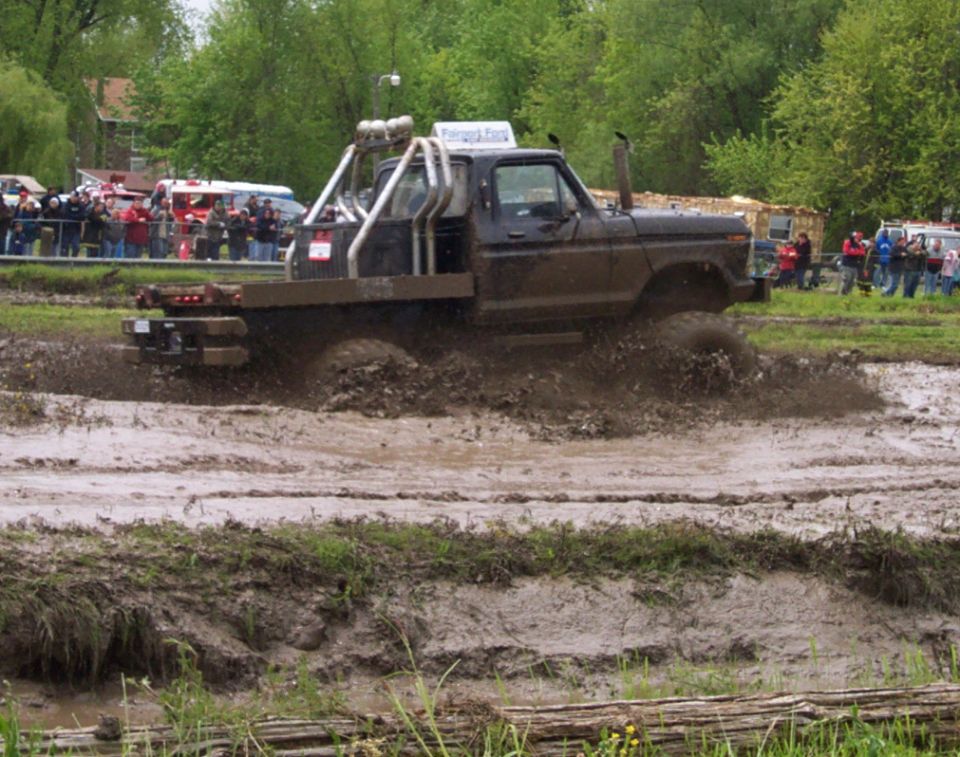Pickup truck with roll cage and flatbed driving through mud. Crowd watches in background