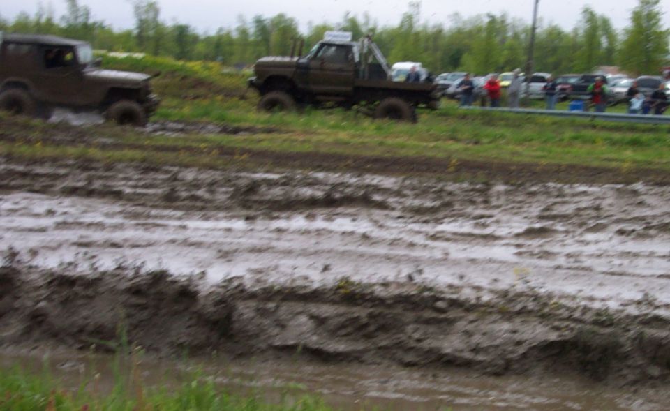 Two muddy off-road vehicles driving through a mud pit with spectators in the background