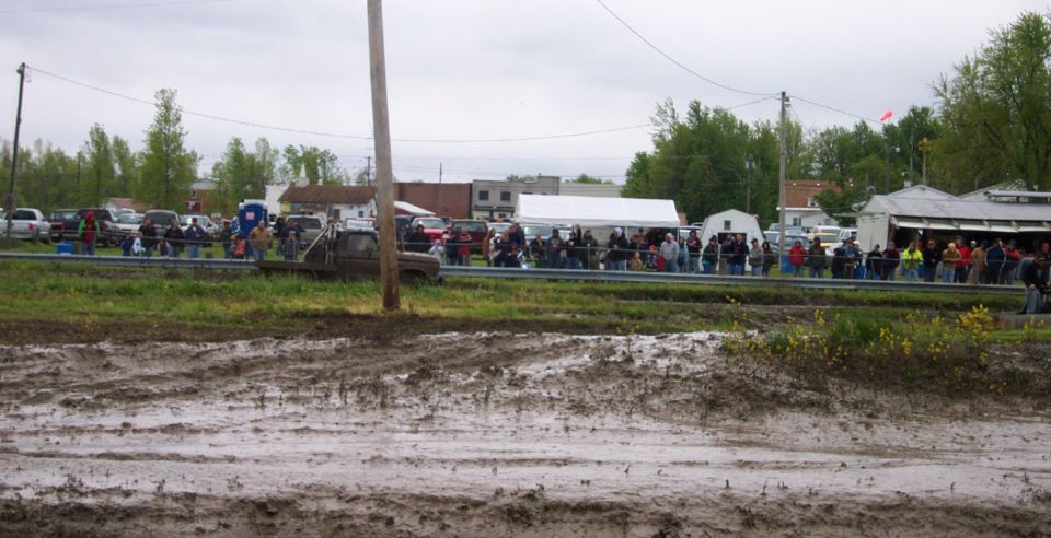 Muddy field with a truck stuck in the mud, spectators watching from a railing, and tents set up