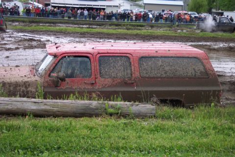 1978 Ford Bronco - A red SUV is stuck deep in a mud pit during an event, with a crowd watching.