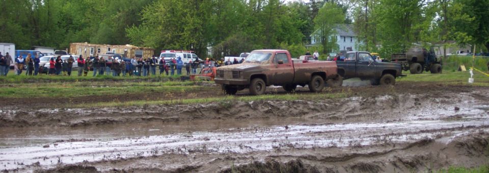 Two pickup trucks driving through a muddy track with spectators in the background