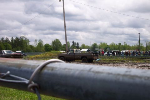 1978 Ford Bronco - A brown pickup truck sits in a muddy field, with a crowd of people and vehicles visible in the distance under a cloudy sky.