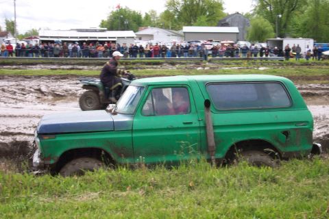 1978 Ford Bronco - A green SUV is stuck in a mud pit during an event, with an ATV passing by and spectators watching.