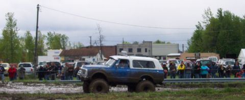 1978 Ford Bronco - A lifted SUV navigates a muddy track while spectators watch from behind a barrier.
