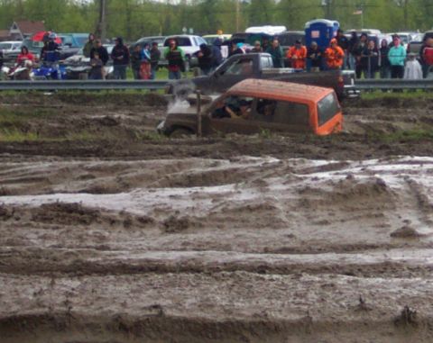 1978 Ford Bronco - An orange SUV is stuck in a mud pit during an event, with onlookers watching from the side.