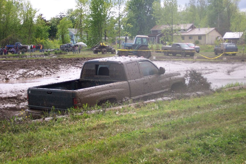 Mud-covered pickup truck stuck in mud, with a tractor and other vehicles in the background
