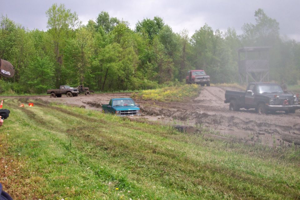 Several trucks are stuck in a muddy off-road course. One teal truck is deep in the mud, while a black truck and a red truck are on a dirt path nearby