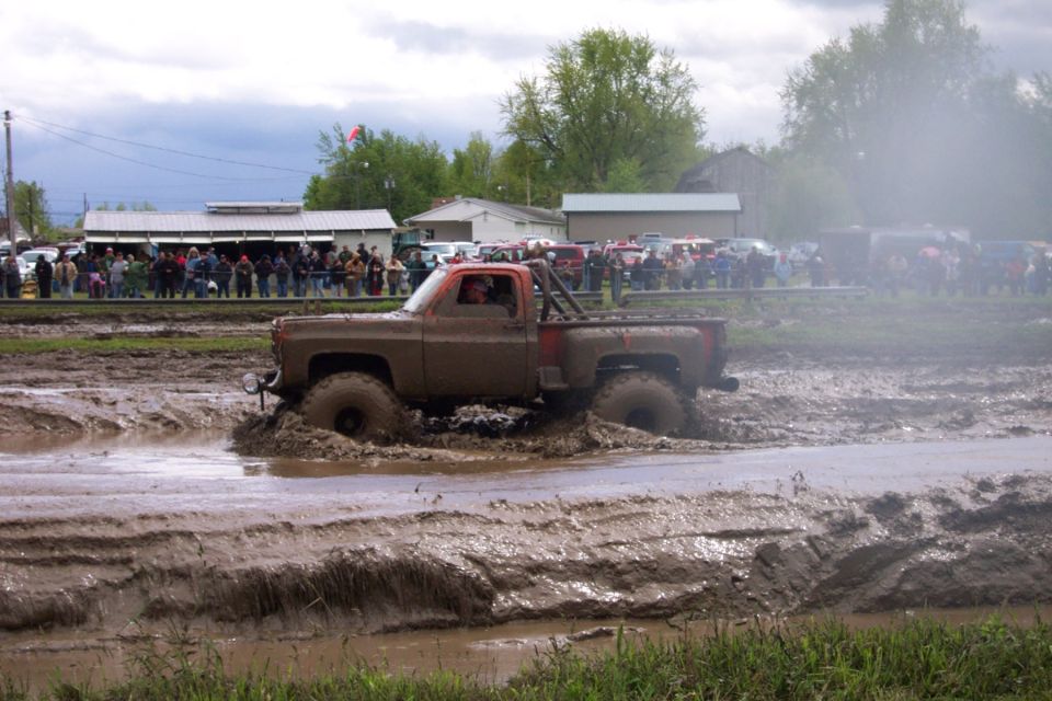 Mud-covered pickup truck with orange interior driving through a mud pit, spectators in background