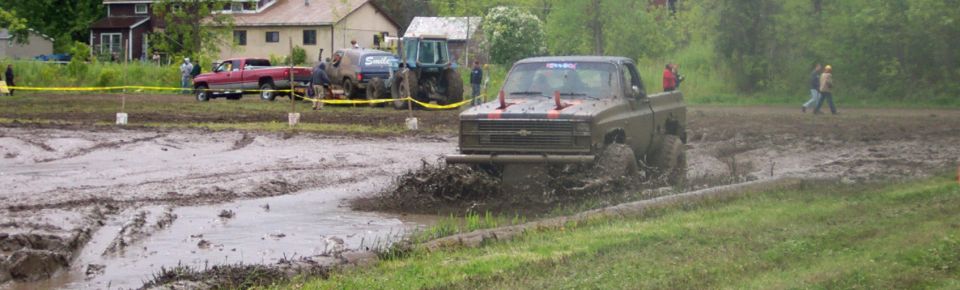 Pickup truck driving through mud, with a red truck and tractor in the background