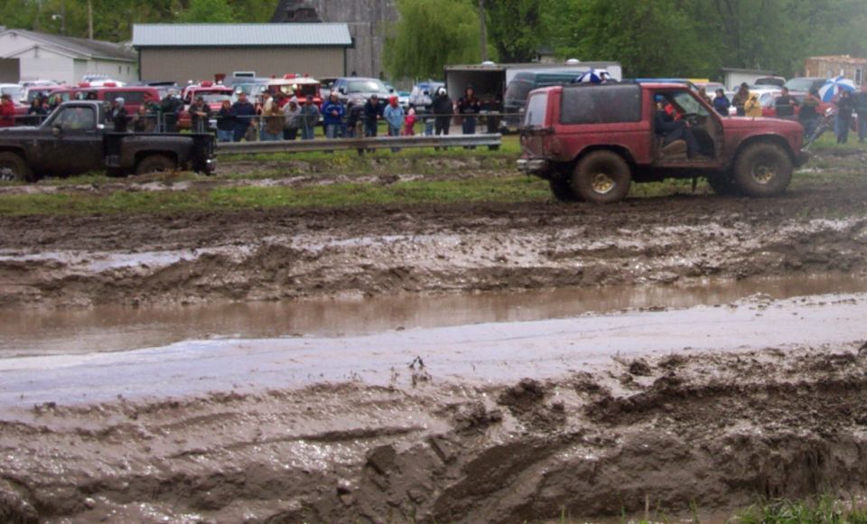Red SUV driving through a muddy track with a black pickup truck stuck in the mud behind it