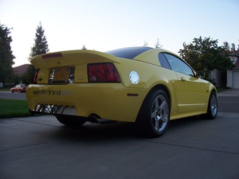 2002 Ford Mustang - This yellow 2002 Ford Mustang features a prominent rear spoiler and dual chrome exhaust tips, viewed from the rear quarter angle.