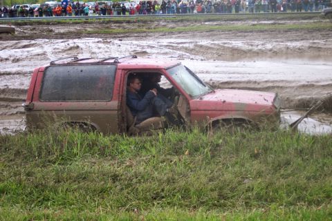 1978 Ford Bronco - A red SUV is stuck deep in the mud, with two people inside and spectators watching in the background.