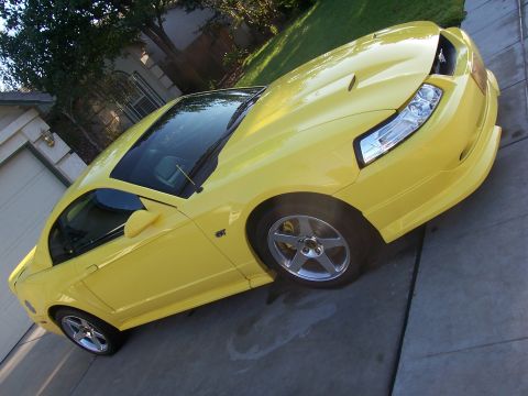 2002 Ford Mustang - This yellow Ford Mustang GT coupe is parked on a driveway in front of a house.