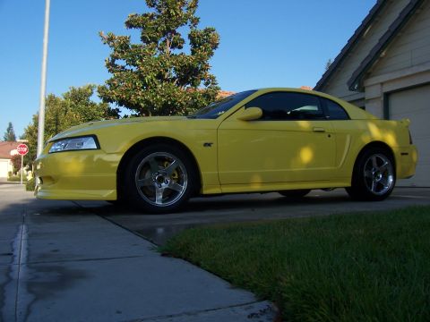 2002 Ford Mustang - A yellow coupe with a GT badge is parked on a driveway under a clear blue sky.
