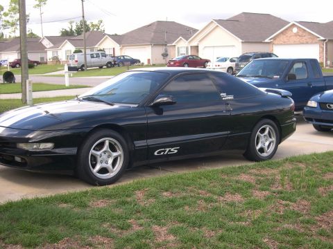 1997 Ford Probe - A black coupe with racing stripes parked in a driveway with other vehicles and houses in the background.