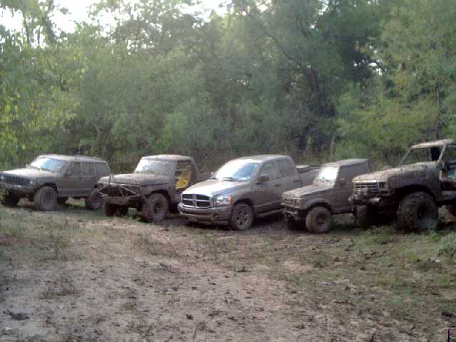 Row of mud-covered 4x4s including a Jeep, Dodge Ram and Ford parked at a wooded trail