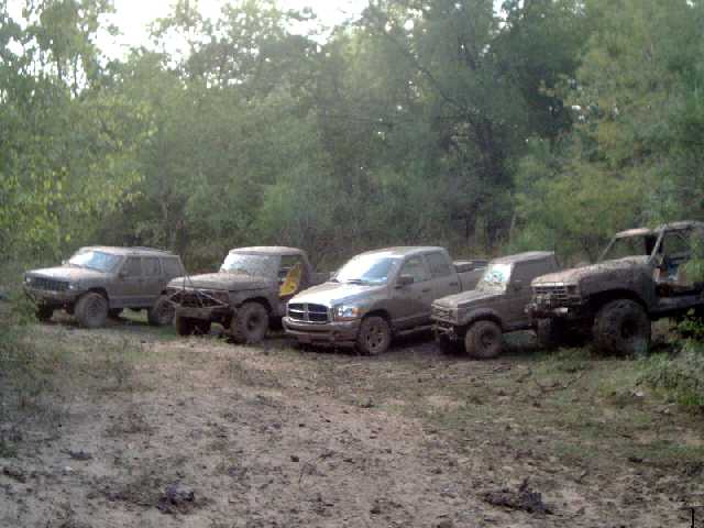 Four mud-covered 4x4s lined up at a wooded off-road trail, including a Jeep Cherokee and a Dodge Ram