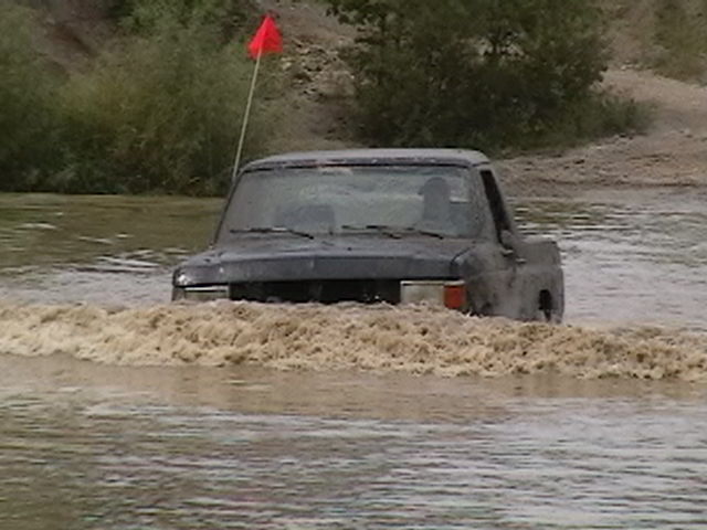 Dark truck with red whip flag crossing deep muddy water, bow wave over the hood