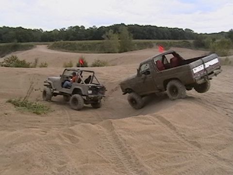 1988 Ford Bronco - Two off-road vehicles navigate sandy terrain with trees in the background.