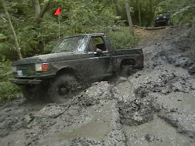 Mud-covered dark pickup truck with a red flag on a pole, stuck in a muddy trail