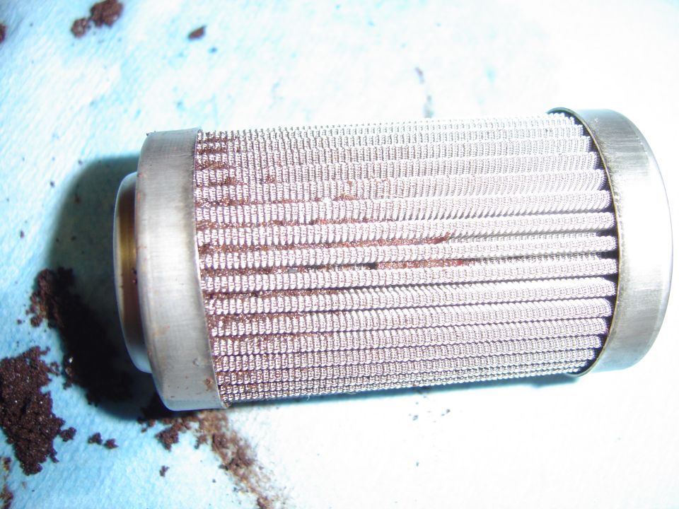 Used fuel filter with metal mesh element and end caps, resting on a blue cloth with debris