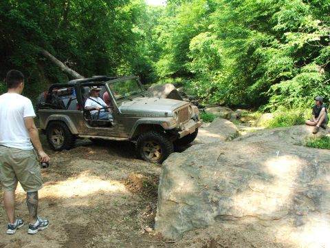 1996 Ford Bronco - A Jeep Wrangler Rubicon is shown off-roading on a dirt path with large rocks and trees.